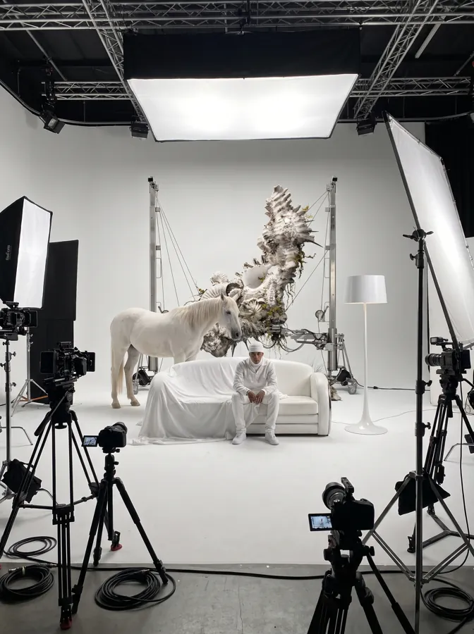 Set de rodaje blanco con un caballo, un sofá y una escultura orgánica suspendida rodeados de cámaras y focos